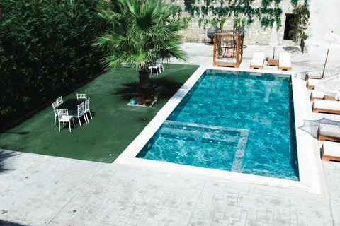 Pool and lawn with a palm tree, white garden chairs and a canopy daybed, enclosed by tall green hedges