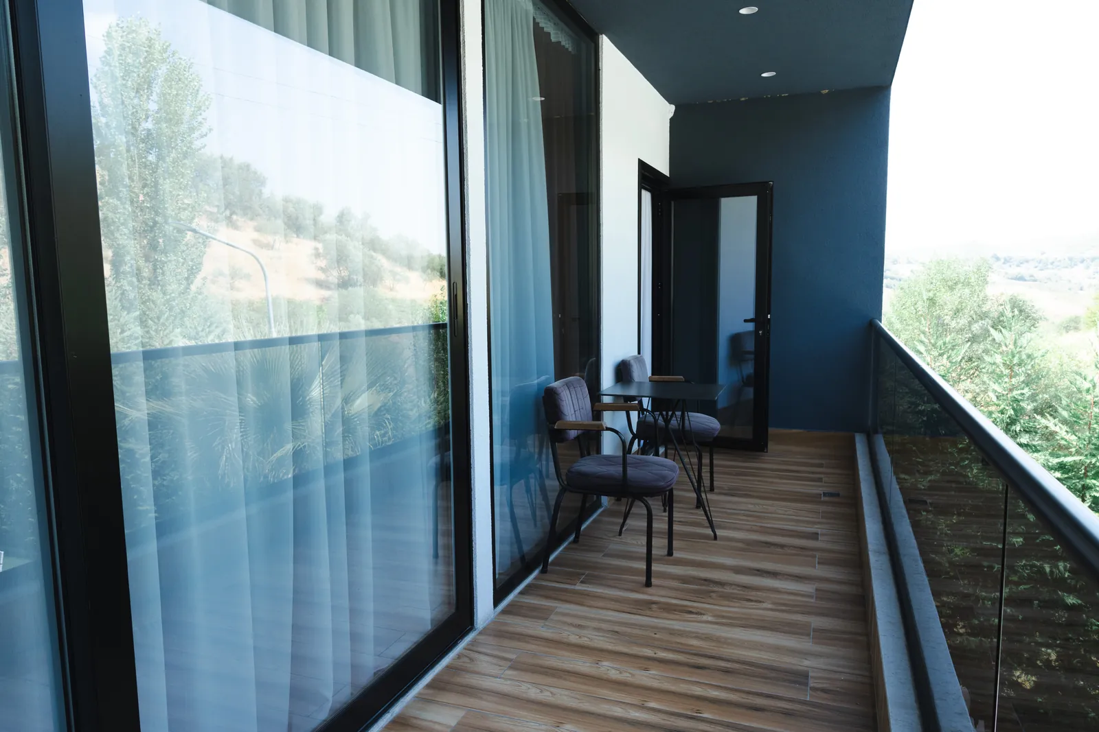 Long balcony with a glass railing and bistro seating, looking out over green hills